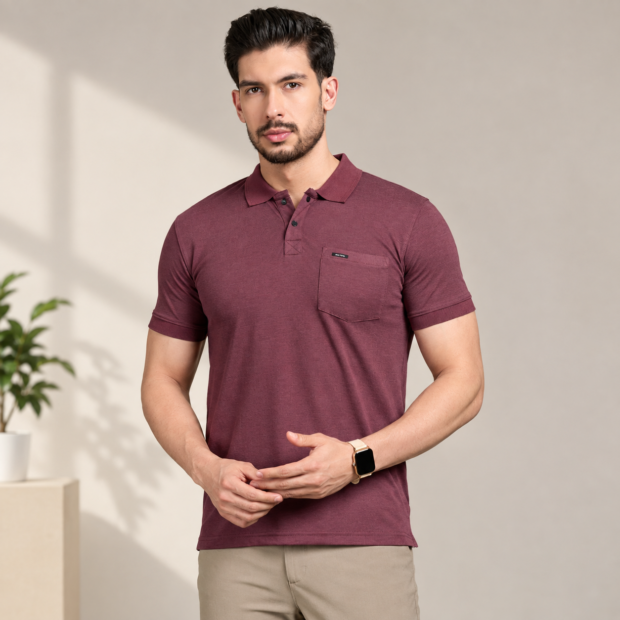 A man wearing a short-sleeve polo shirt in maroon with a chest pocket, paired with beige trousers, standing in a studio setting with soft shadows.